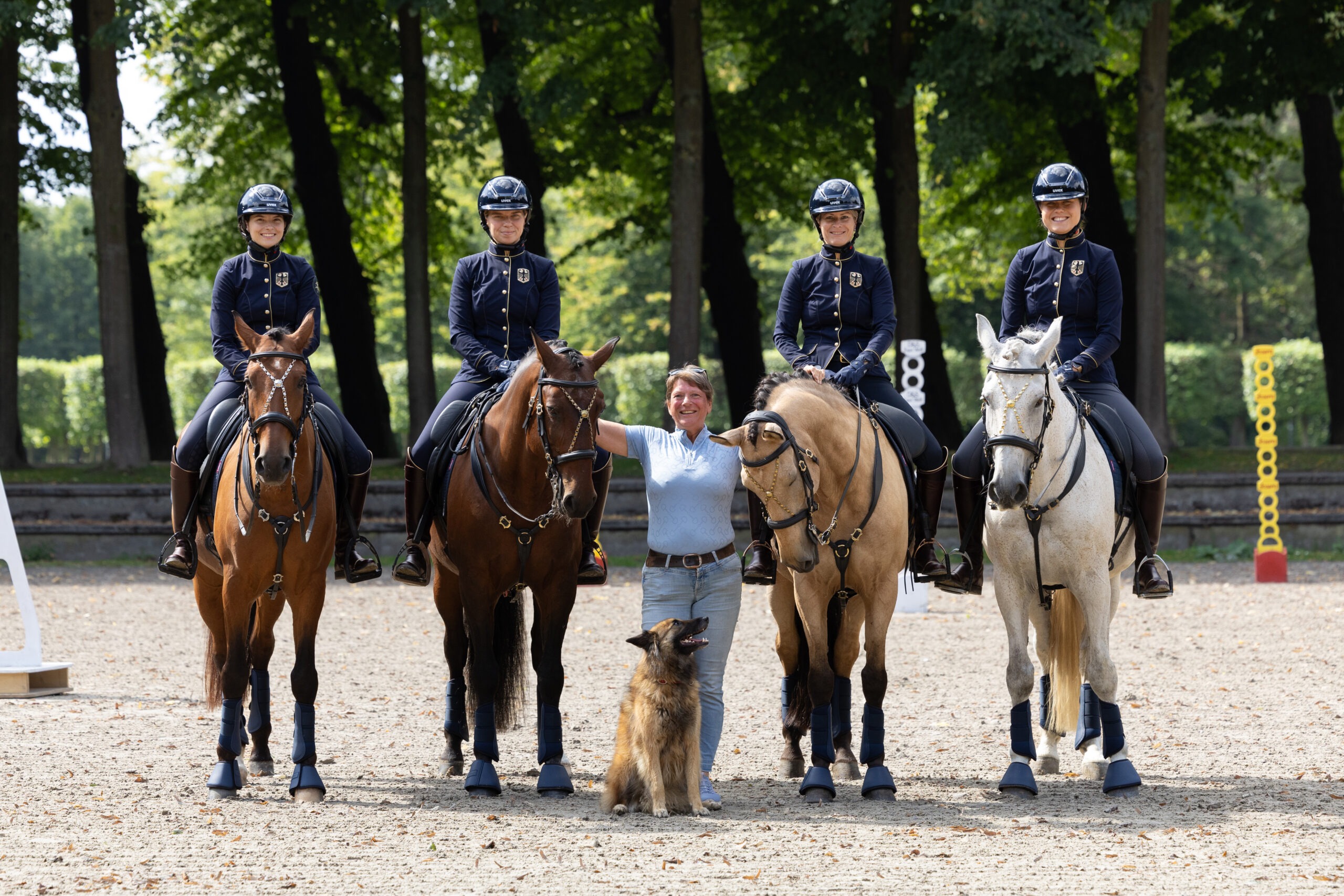 Vier deutsche Working-Equitation-Reiterinnen in dunkelblauer Reitbekleidung sitzen auf ihren Pferden. Gemeinsam mit einer Frau stehen sie auf einem Reitplatz vor einer bewaldeten Kulisse und präsentieren sich als deutsches Team.